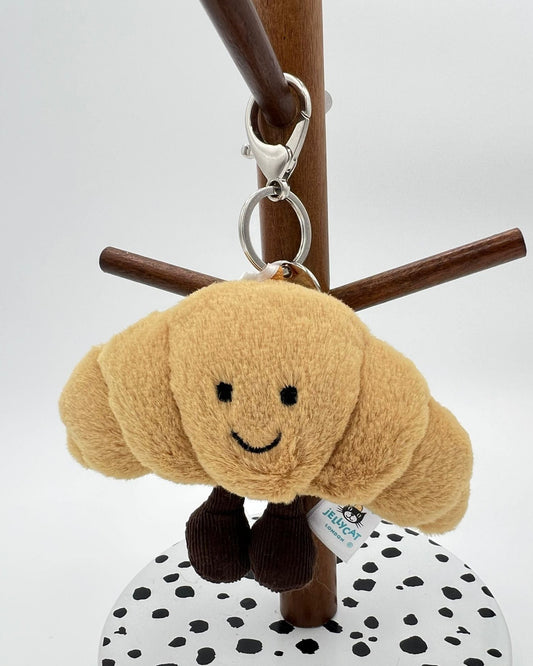 Plush croissant-shaped keychain with a smiling face on a stand against a white background